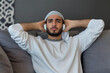 Dissatisfied and angry young Arabian man sitting on sofa at home and covering ears from excessive noise