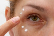 Close up cropped eye of middle aged caucasian woman looking at camera applying cream on skin