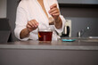 Close-up woman in white bathrobe, sweetening his tea, standing at kitchen table with open digital tablet, in a minimalist home kitchen interior