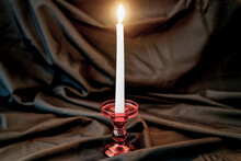 White Candle, Fire. Red Glass Candlestick. Dark Fabric Background.