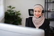 Call center worker, young Muslim woman wearing hijab, talking to customer on call phone on computer in customer service office