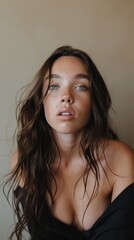 Wall Mural - A woman in a black top, with long wavy hair and blue eyes poses for a portrait against a beige wall and looks straight into the camera.