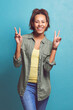 Happy woman, portrait and peace sign with fashion for style, love or care on a blue studio background. Female person, brunette or model with smile, emoji or calm attitude in cool casual clothing