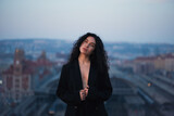 Fototapeta Zwierzęta - Attractive young curly brunette woman in a black jacket and black pants stands on a rooftop against a historic building during a magical sunset.