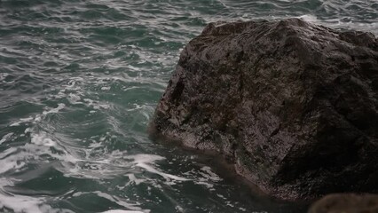 Poster - A large rock is in the ocean with waves crashing around it. The water is a deep blue color and the rock is a dark grey color. Scene is calm and peaceful, as the rock stands strong against the waves
