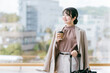 © buritora - 駅・ターミナルのエントランスでコーヒーを飲むビジネスウーマン（待ち合わせ・デート・待つ・温まる）