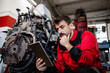 Serviceman ordering spare parts on tablet computer and repairing tractor engine.