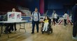 © Framestock - Woman with SMA in electric wheelchair comes with polling officer to voting booth. African American woman gives vote controller to voter. National Election Day in United States. Concept of civic duty.