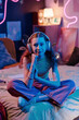 Vertical shot of stylish teen girl wearing headphones sitting on bed in her bedroom decorated with neon light late in evening
