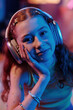 Vertical closeup portrait of cheerful Caucasian girl with red hair wearing headphones looking at camera, neon lighting