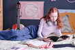Modern Caucasian schoolgirl relaxing on bed in her bedroom looking through notes in notebook, copy space