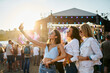 © artiemedvedev - Group of young women take selfie at summer beach music festival. Happy friends party, dance in crowd. Sun sets behind stage. Girls enjoy live concert, fun vacation. Casual fashion, youth culture.