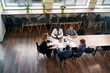 © artiemedvedev - Top view of a diverse group of professionals in a meeting. A female CEO leads a business consultation at a modern office table with multiethnic teammates discussing strategy, growth, and plans.