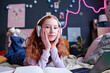 Portrait of modern teen schoolgirl with red hair wearing headphones posing for camera while doing homework in her bedroom