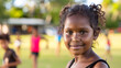 an aboriginal girl wearing athletic wear. she is facing the camera and standing on school ground with her friends playing footy in the background 