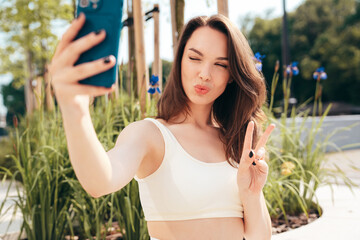 Wall Mural - Young beautiful smiling female in trendy summer white cycling shorts and tank top clothes. Sexy carefree woman posing in street. Positive model.  Slim and fit body. In sunglasses. Takes selfie photos