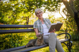 Fototapeta Na drzwi - Happy senior woman enjoys reading book and spending time with her cat while sitting on a bench in her garden.	