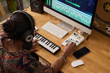 Young male musician listening to new music in headphones while sitting by desk in front of computer and turning console to adjust sounds