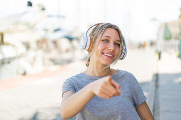 Poster - Young blonde woman at outdoors listening music and pointing to the front