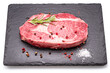Raw ribeye steak with pepper corns and rosemary on graphite serving board isolated on white background.