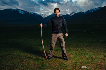 Poster - Handsome man hiker with stick standing on mountains background at dusk