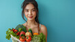 Indoor shot of cheerful dark haired Asian woman holds freshly made smoothie from vegetables carries net bag wears cropped top and leggings isolated over blue background blank space for promo.
