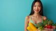 Indoor shot of cheerful dark haired Asian woman holds freshly made smoothie from vegetables carries net bag wears cropped top and leggings isolated over blue background blank space for promo.