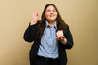 Pretty Latin overweight businesswoman posing with piggy bank and making ok sign in a studio setting