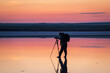 Serene scene with a photographer silhouetted against a vibrant sunset, standing in calm waters while setting up a camera on a tripod.