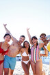 Cheerful portrait of five diverse friends on the beach having fun while posing and looking at the camera smiling and raising their arms. Young multiethnic people in swimsuit on summer holidays.