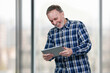 © DenisProduction.com - Smiling mature man holding tablet pc in both hands. Huge office windows background.