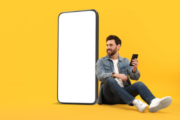 Poster - Man with mobile phone sitting near huge device with empty screen on orange background. Mockup for design