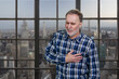 Portrait of a mature man having heartache touching his chest. Aged man having heart atack indoors. Checkered windows background with cityscape view.