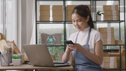 Go green asian SME owner chat online happy talk AI chatbot phone. Young seller asia woman typing mobile work at home office desk easy selling smart CRM platform app for eco shop retail net zero store.