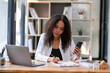 © Wasana - Cheerful professional woman waving to the smartphone during a friendly video call in a sunny office setting.