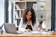© Wasana - Cheerful professional woman waving to the smartphone during a friendly video call in a sunny office setting.