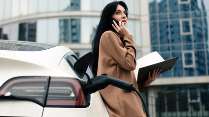 A businesswoman waits for her electric car to charge in the parking lot of a business center. Futuristic eco-friendly electric car uses alternative energy in urban lifestyle.