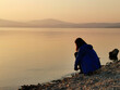 Female looking sunset on sea, lake. Woman sitting on rocks. Lonely person. Girl alone outside. Vacation on sea. Relax sunset silhouette. Girl from behind.