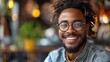 A cheerful young man with dreadlocks and glasses smiling confidently in a casual indoor setting. 