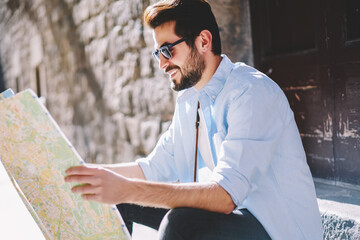 Wall Mural - Smiling bearded traveler in spectacles reading map to find right direction of showplaces sitting outdoors on street.Positive young man tourist in sunglasses search destination during trip in old town