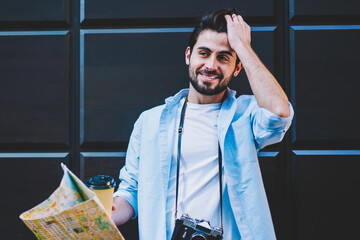 Wall Mural - Cheerful bearded male tourist touching hair on black promotional background with copy space area.Handsome traveler with vintage camera holding map in hand while standing outdoors on street