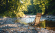 © visoot - In a sunny outdoor camping, there is a small stream behind and an outdoor folding chair on a small gravel road surface. Generative AI.