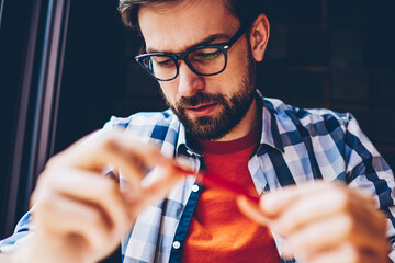 Wall Mural - Pensive bearded young man in optical eyeglasses for vision correction studying in coworking space.Concentrated hipster guy dressed in casual stylish shirt sitting in coffee shop interior