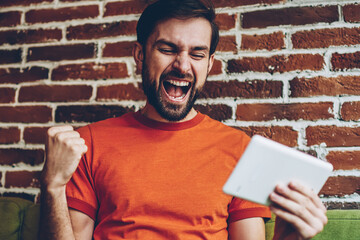 Wall Mural - Overjoyed bearded young man celebrating victory of favourite team watching live stream online on website on modern tablet device connected to 4G internet.Excited hipster blogger got win on web page