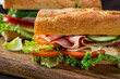 Two big sandwich with lettuce, tomatoes prosciutto cucumber and cheese on a wooden cutting board on a dark background