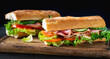 Two big sandwich with lettuce, tomatoes prosciutto cucumber and cheese on a wooden cutting board on a dark background