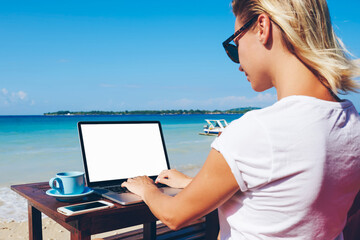 Wall Mural - Young woman millennial keyboarding on netbook with mock up screen communicating in online chat sitting near ocean, businesswoman working distantly on vacation in tropical environment enjoying nature