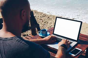 Fototapete - Back view of skilled male freelancer doing distance job on ocean shore browsing information on laptop computer,man travel blogger typing post for update content on website recreating on sea beach