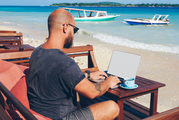 Fototapete - Skilled male freelancer working online on his business via laptop computer while resting on ocean shore, millennial hipster guy update content in his blogg in keyboarding publication about trip