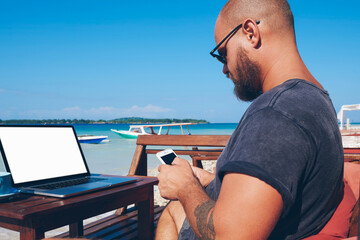 Fototapete - Young man using modern technology and 4G internet working remotely on ocean beach in summer, millennial male blogger checking notification on smartphone update his web page content on journey